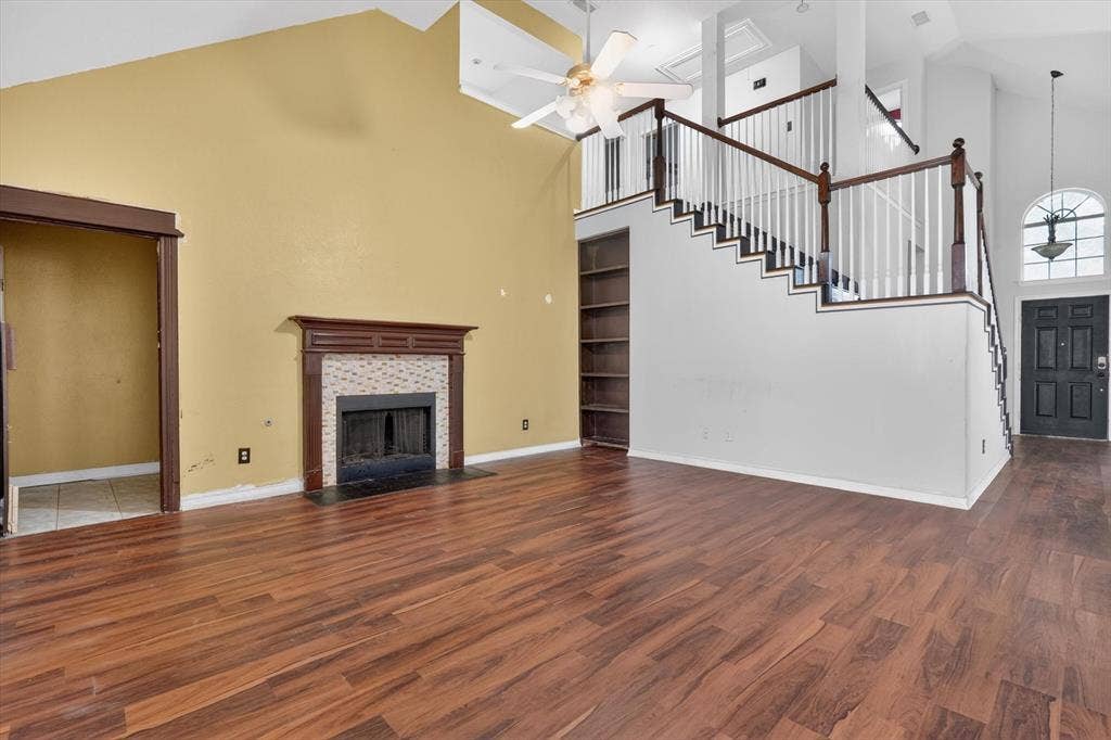 Living area featuring vaulted ceilings, wood-finish flooring, a fireplace with a decorative mantel and tiled surround, a multi-level staircase with white balusters and dark wood handrails, and a built-in bookshelf