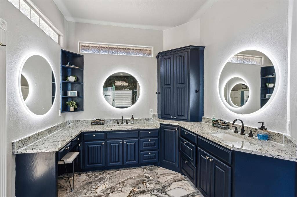 2100 Gene Court, Granbury, TX 76048 Dual backlit circular mirrors highlight the expansive vanity featuring dark blue cabinetry, light granite-finish countertops, and two undermount sinks
