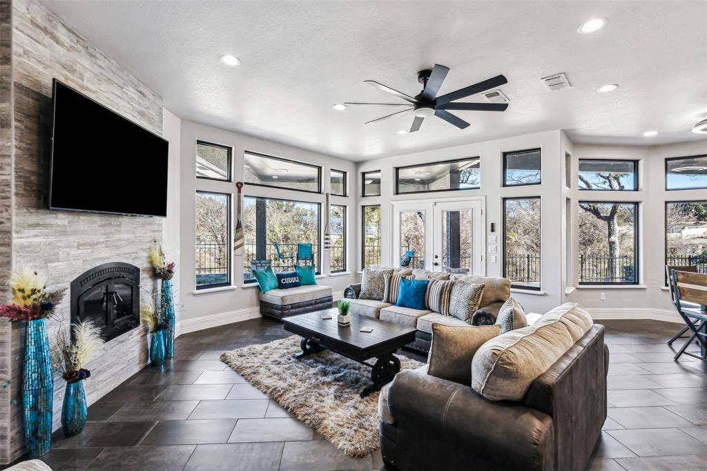 2100 Gene Court, Granbury, TX 76048 Spacious living area featuring a stacked stone fireplace with an arched firebox, dark tile flooring, a multi-pane glass door opening to an exterior area, and extensive picture windows