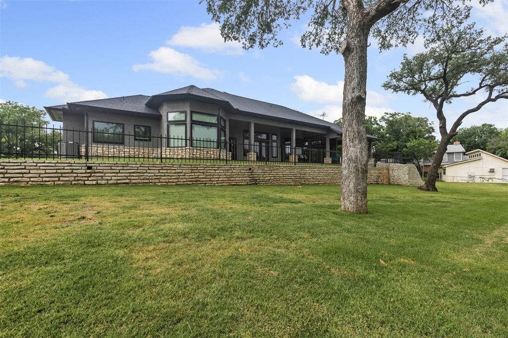 2100 Gene Court, Granbury, TX 76048 Contemporary stucco exterior with a dark gray roof, multiple large windows, a stone retaining wall, and a covered patio area