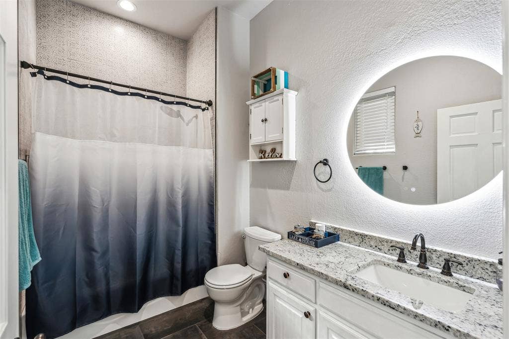 2100 Gene Court, Granbury, TX 76048 Bathroom featuring a built-in shower with tiled walls, a single vanity with a light-colored countertop, white cabinetry, a built-in toilet, and an illuminated circular mirror
