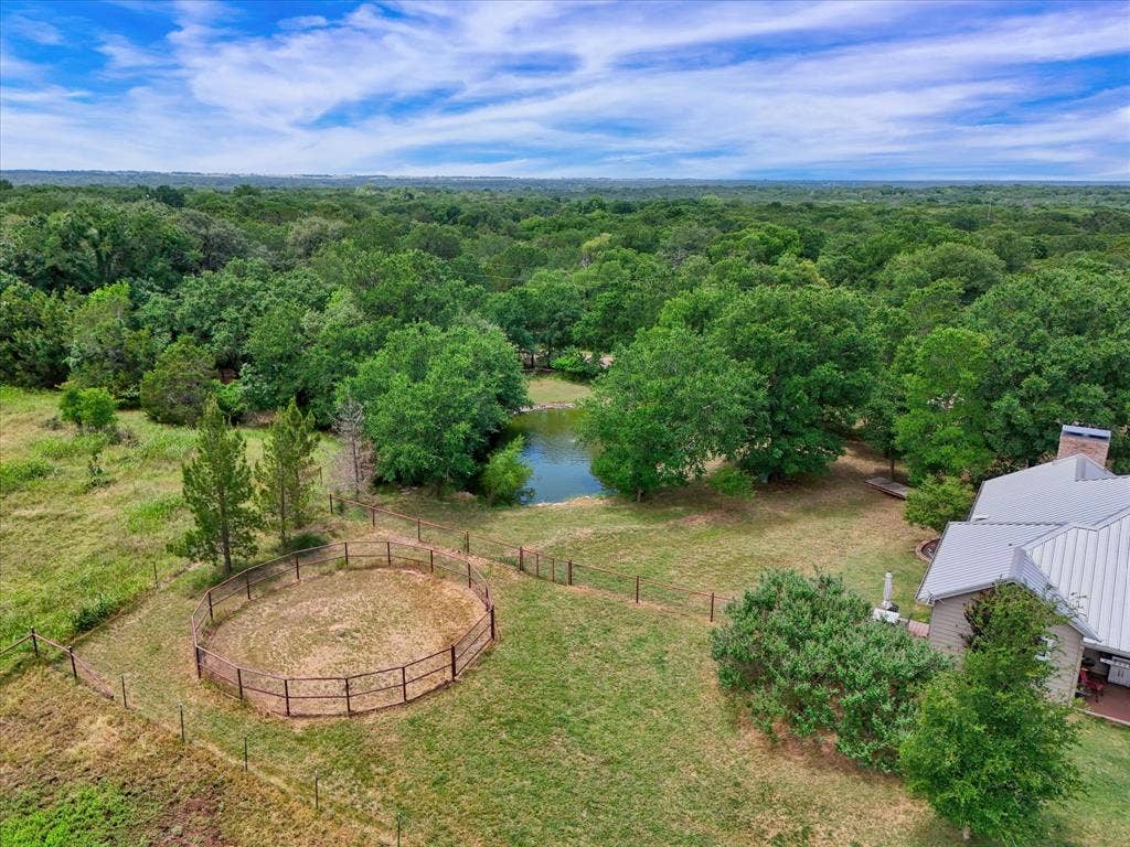 613 Bone Road, Valley Mills, TX 76689 Expansive property featuring a tranquil pond surrounded by mature trees, a fenced round pen, and a residence with a metal roof and brick chimney