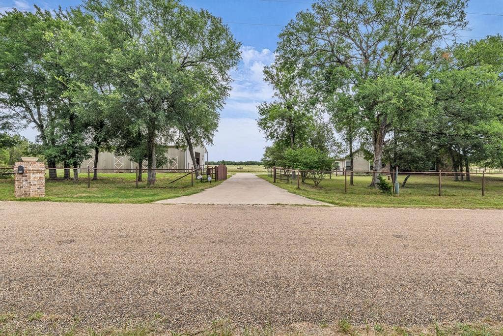 613 Bone Road, Valley Mills, TX 76689 Paved driveway entry framed by mature trees, leading to a property featuring a brick mailbox column, perimeter fencing, and a barn structure