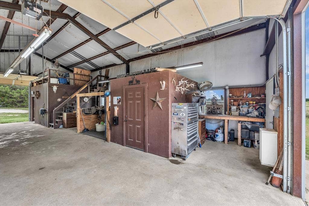 613 Bone Road, Valley Mills, TX 76689 Spacious utility building featuring a concrete floor, exposed metal beam structure, multiple fluorescent light fixtures, a roll-up garage door, and a built-in workbench with shelving