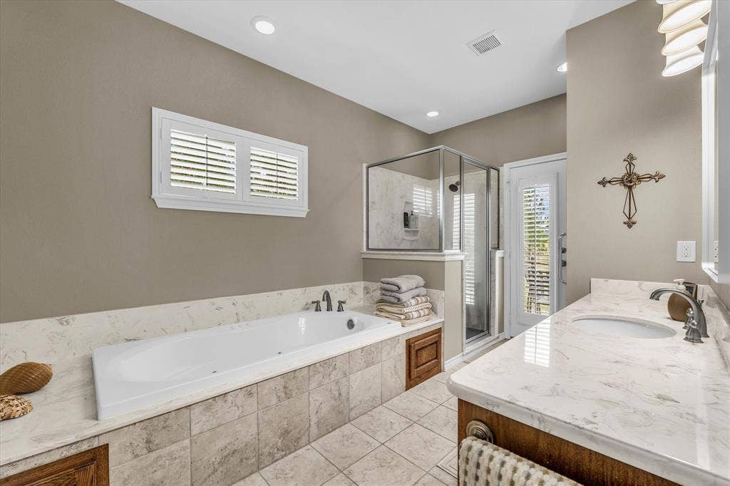 613 Bone Road, Valley Mills, TX 76689 Bathroom featuring a built-in soaking tub, a glass-enclosed shower, a double vanity with undermount sinks, and a large window with plantation shutters