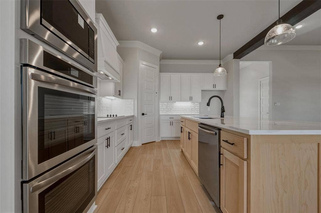 2305 Quartz Street, Krum, TX 76249 Stainless steel wall oven and microwave, white shaker cabinetry with black hardware, wood-finish flooring, light-toned kitchen island with a built-in sink, and pendant lighting