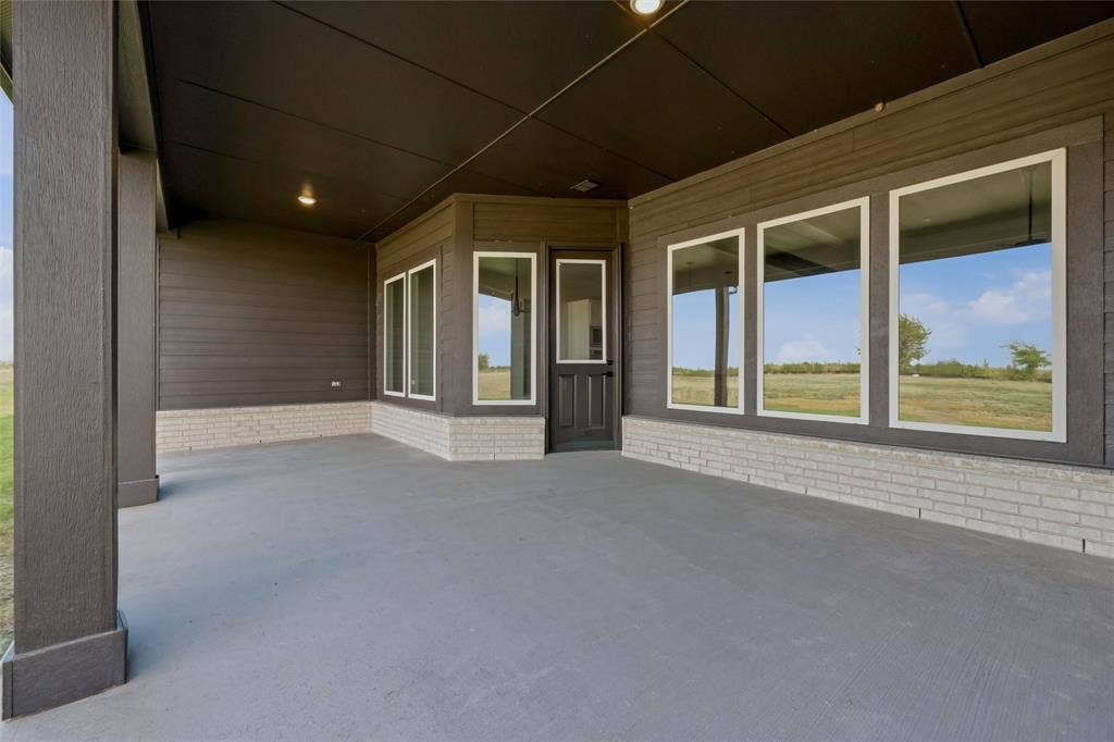 2305 Quartz Street, Krum, TX 76249 Spacious covered patio with a textured concrete surface, framed by a dark plank siding exterior and a light brick wainscoting