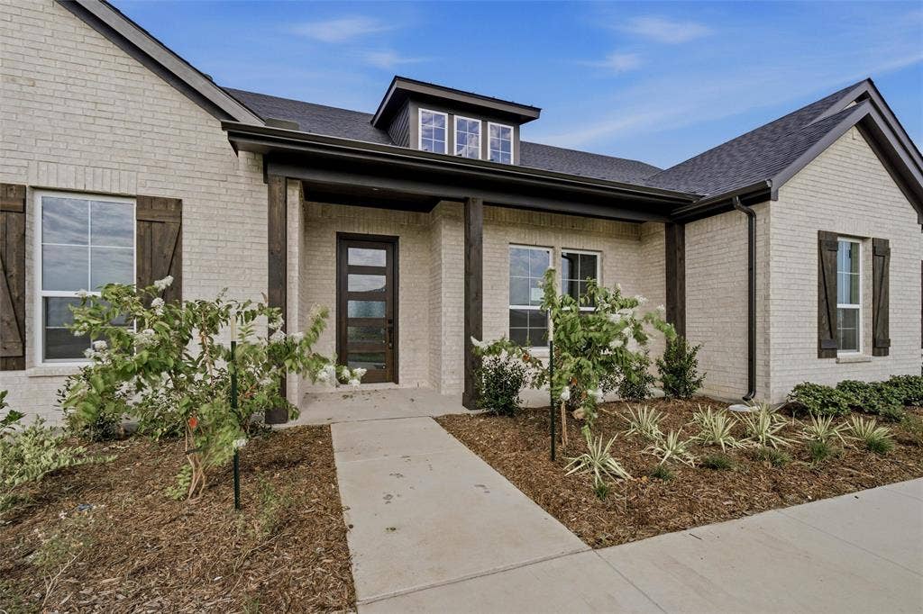 2305 Quartz Street, Krum, TX 76249 Light-colored brick exterior featuring dark trim, a covered entry with wood-wrapped columns, and a dark asphalt shingle roof