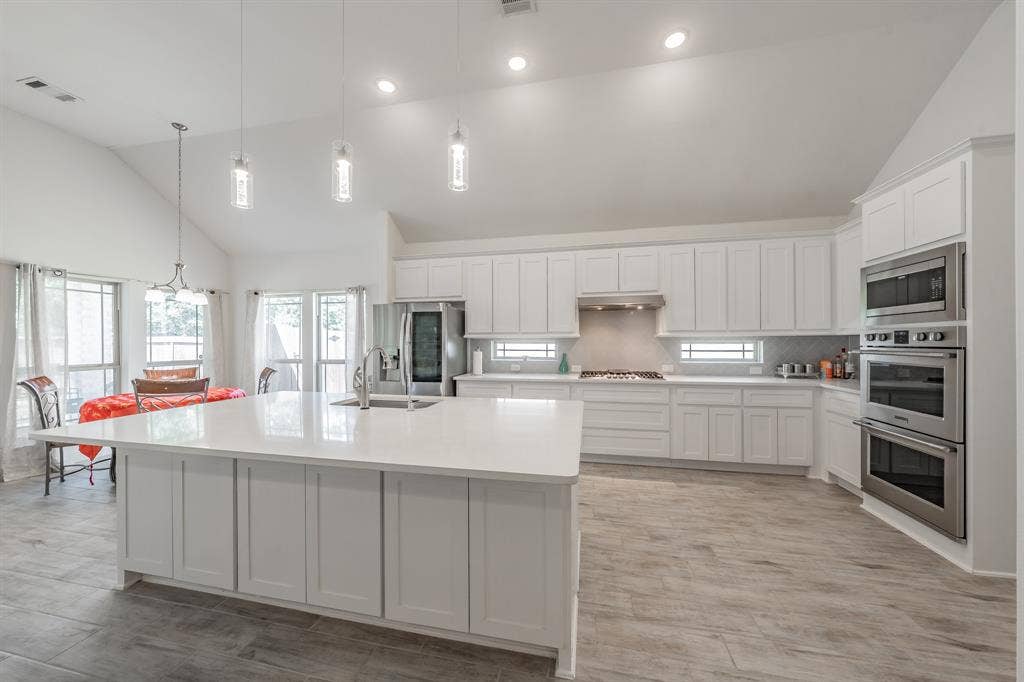Kitchen with decorative light fixtures, stainless steel appliances, white cabinetry, an island with sink, and high vaulted ceiling