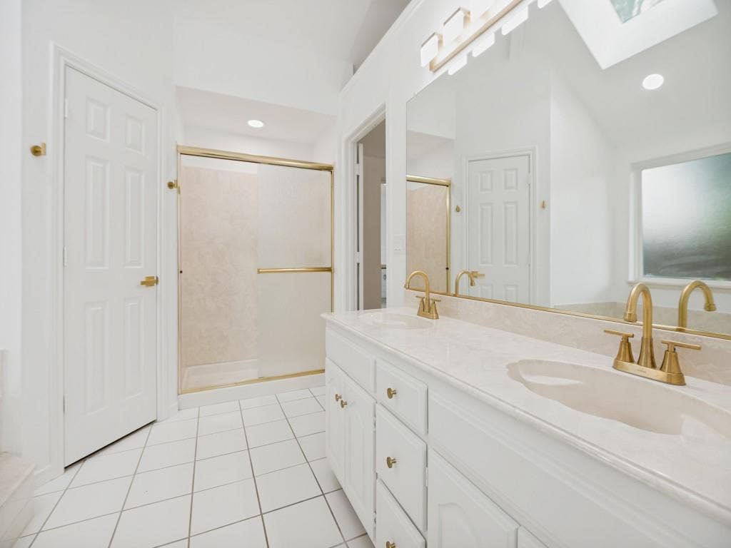 4312 Avonshire Lane, Plano, TX 75093 Bathroom featuring a dual vanity with integrated sinks and brass fixtures, a large mirror, a shower with a brass-framed enclosure, white tile flooring, and a skylight