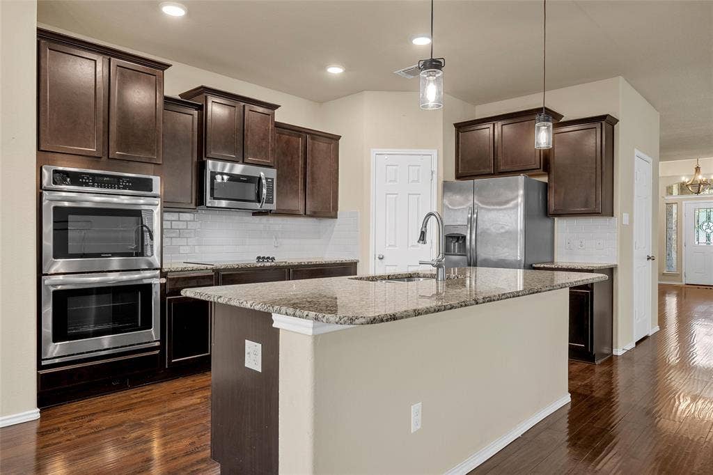 Kitchen featuring dark wood cabinetry, stainless steel double ovens, a built-in microwave, white subway tile backsplash, and a large island with a stone countertop and an undermount sink