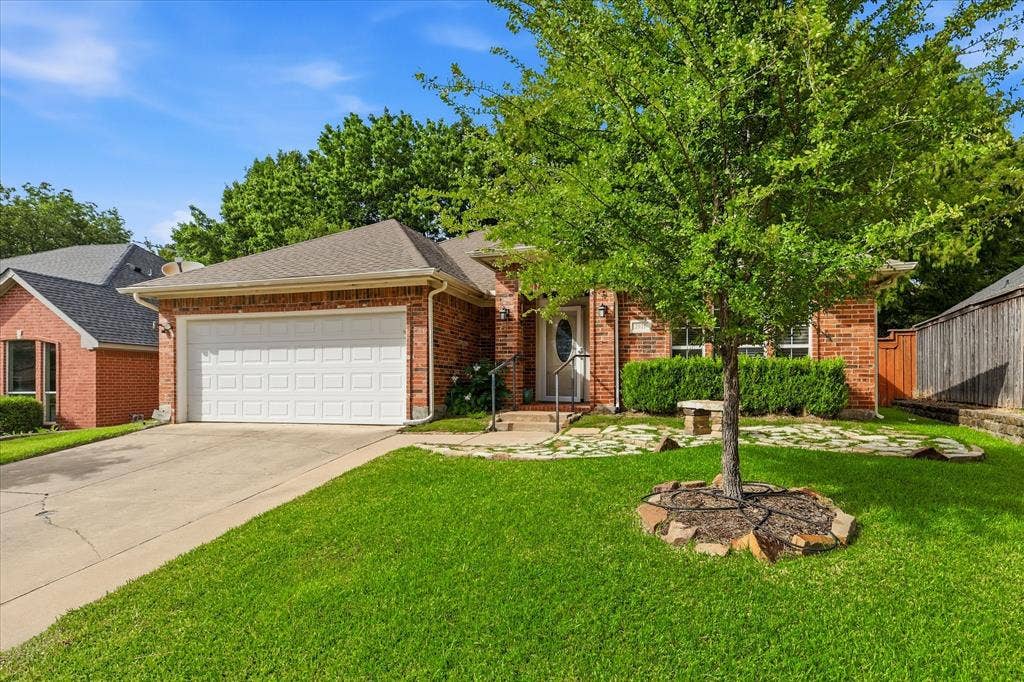 Brick exterior featuring an attached two-car garage, mature front yard tree, and a stone-lined garden bed