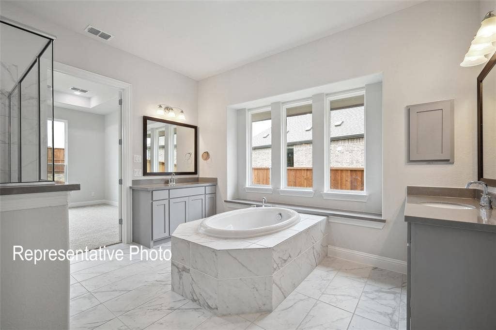 2297 Slate Drive, Waxahachie, TX 75167 Spacious bathroom featuring a built-in soaking tub with marble-look tile surround, dual vanities with integrated sinks and quartz-look countertops, a separate glass-enclosed shower, and large windows above the tub