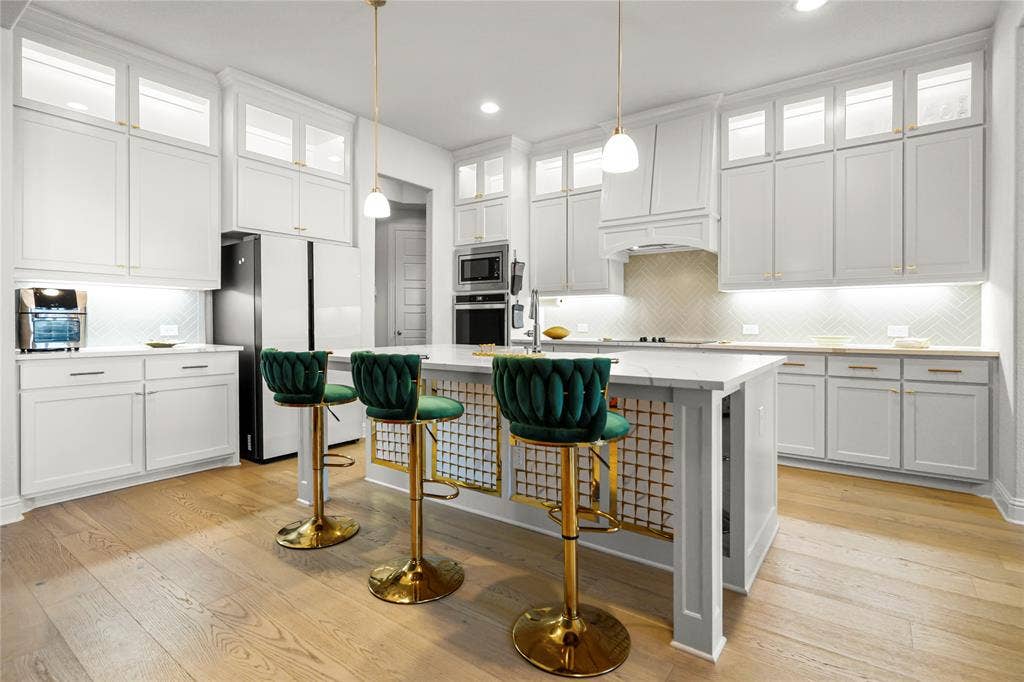 Contemporary kitchen featuring light wood-finish flooring, white shaker-style cabinetry with gold-tone hardware, a light-toned herringbone tile backsplash, and a central island with a light countertop