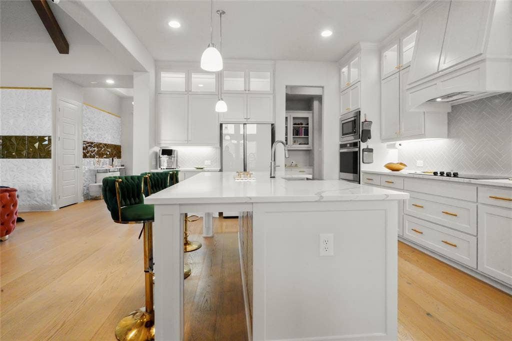Open-concept kitchen featuring an expansive central island with a solid surface countertop, white cabinetry with gold-finish hardware, built-in stainless steel appliances, and wood-finish flooring