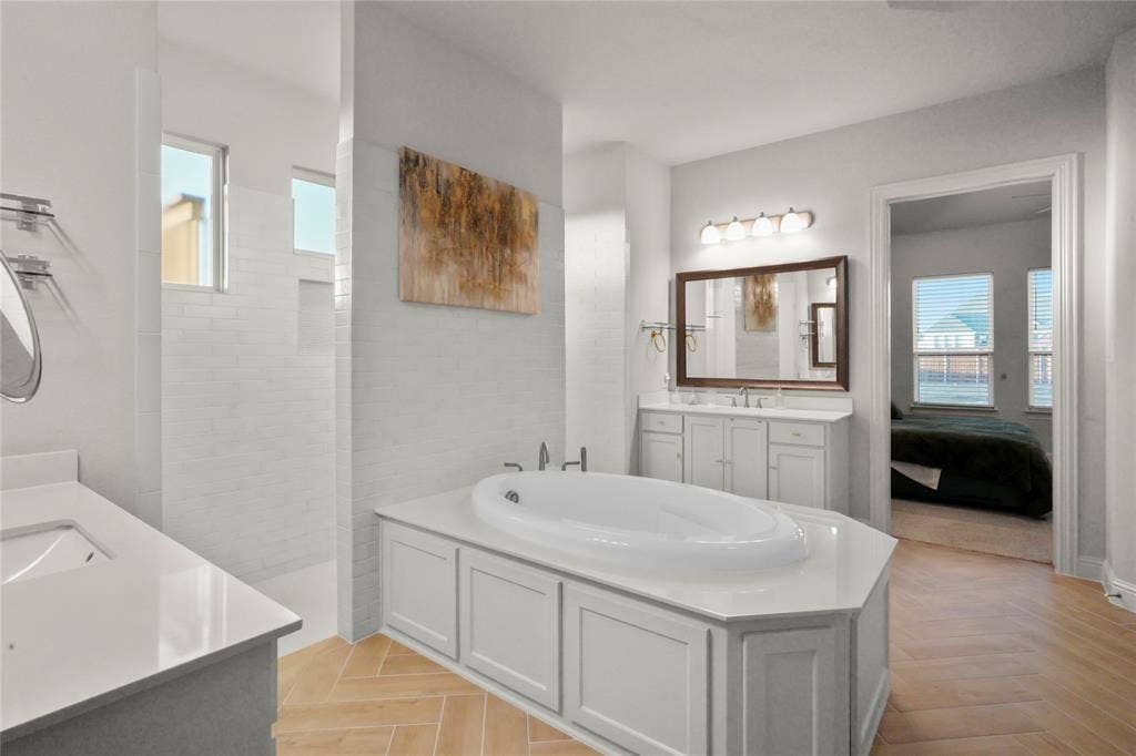 Expansive bathroom featuring a built-in soaking tub, dual vanity with quartz countertops, wood-finish flooring in a herringbone pattern, and a walk-in shower with white subway tile