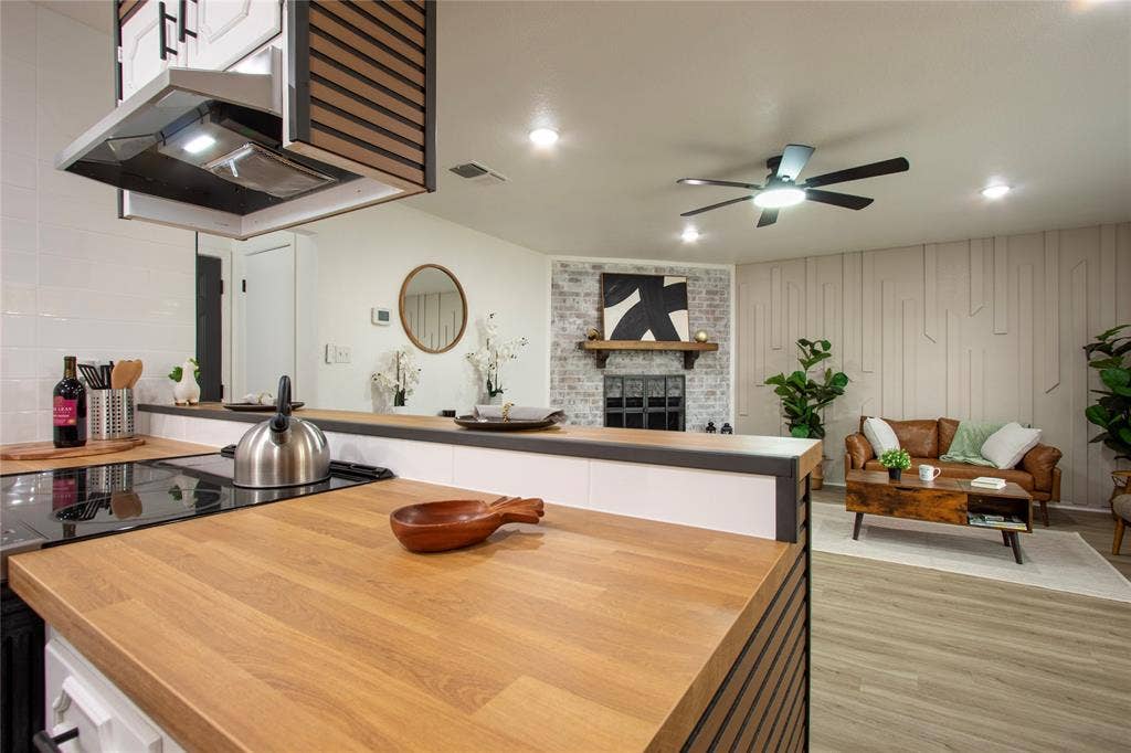 1205 Augusta Road, Benbrook, TX 76126 Open-concept design featuring wood-finish countertops, a modern range hood with integrated lighting, a built-in electric cooktop, light wood-finish flooring, and a brick-surround fireplace with a wood mantle