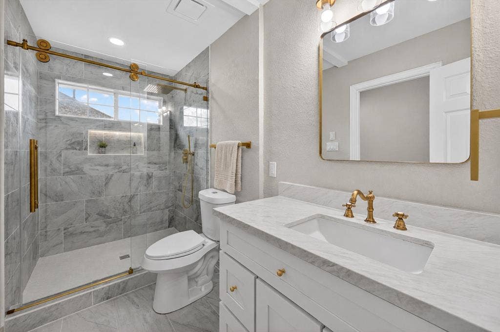 4437 Lone Tree Drive, Plano, TX 75093 Bathroom featuring a glass-enclosed shower with gold-tone hardware, a tiled shower surround with a recessed niche, a white vanity with a rectangular undermount sink, a stone-finish countertop, and gold-tone vanity faucet