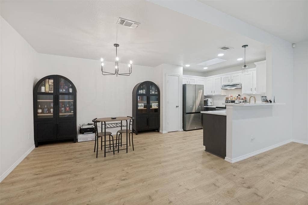 Open-concept living area with light wood-finish flooring, a modern chandelier, a kitchen with white cabinetry, stainless steel appliances, and a central island with a breakfast bar