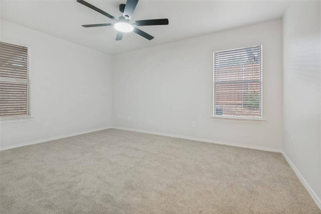 Spacious room featuring light-colored carpeting, two windows with blinds, a ceiling-mounted fan with integrated lighting, and neutral wall finishes