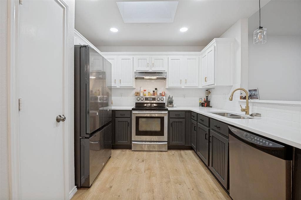 Contemporary kitchen featuring two-tone cabinetry, stainless steel appliances, light-toned wood-finish flooring, recessed lighting, and a skylight