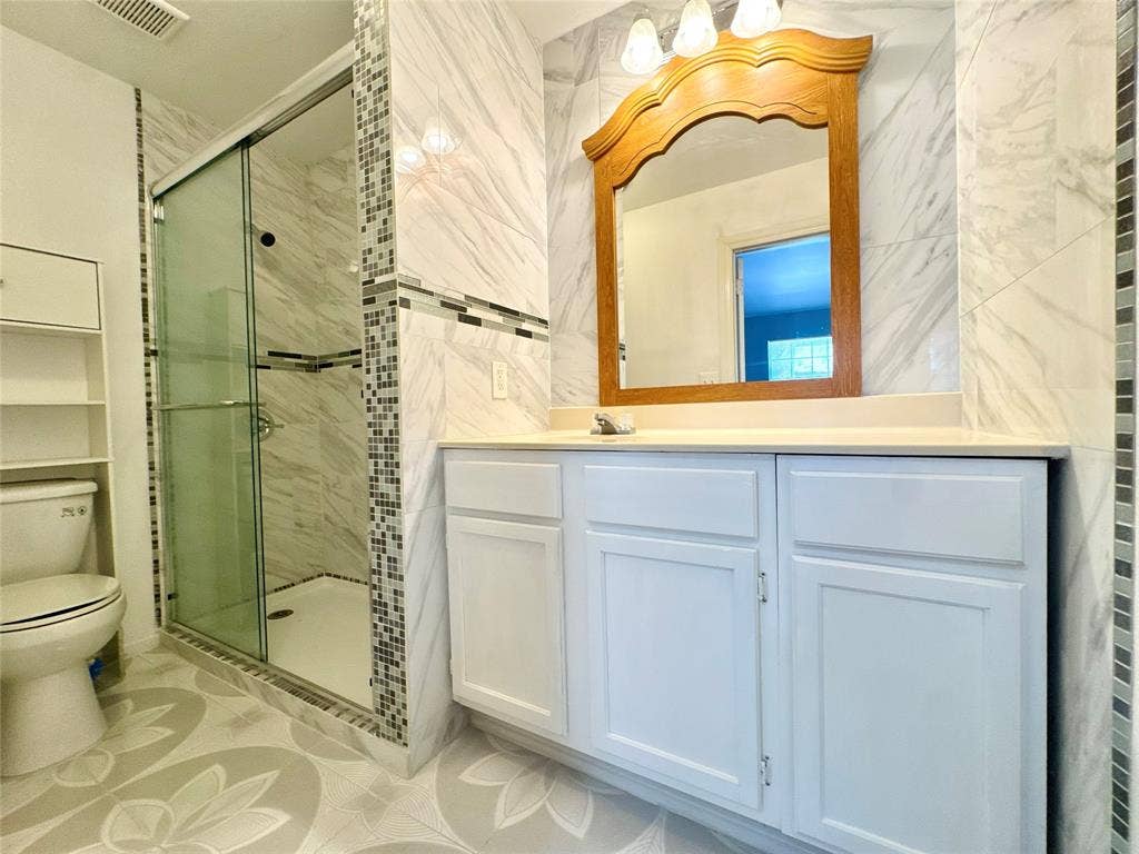 Bathroom featuring a glass-enclosed shower with tile surround, a built-in vanity with white cabinetry, a light-colored countertop, and a decorative mirror with an integrated light fixture