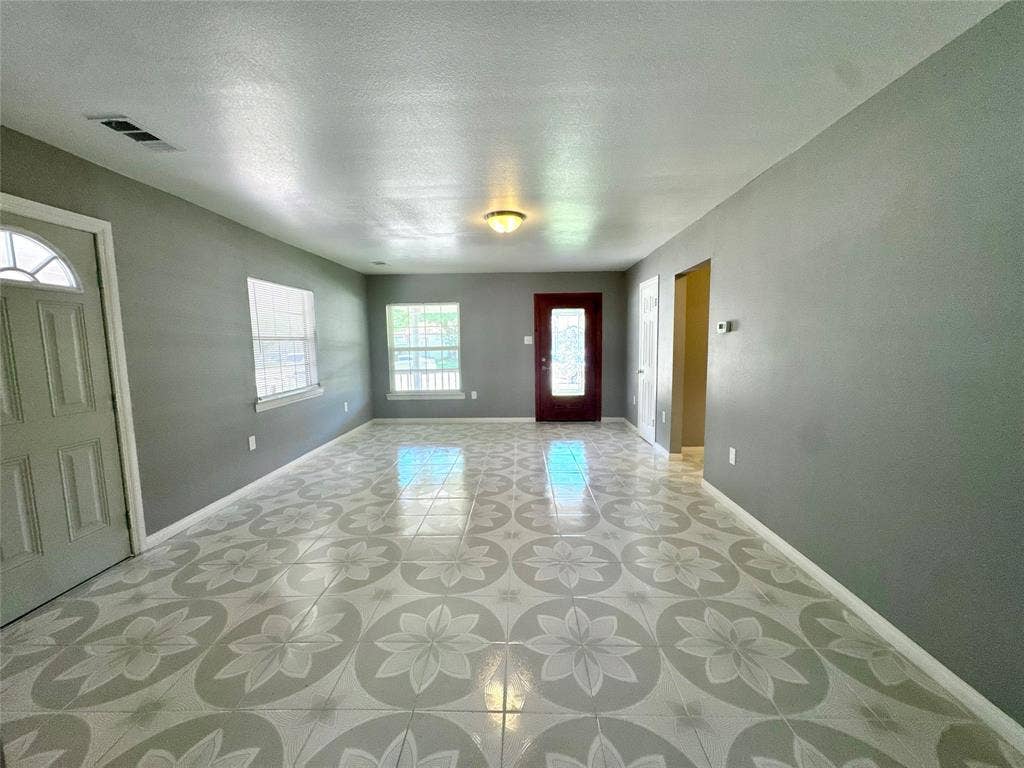 Spacious room featuring patterned tile flooring, a semi-flush mount ceiling light, two windows, a paneled entry door with an arched glass insert, and a dark-toned interior door