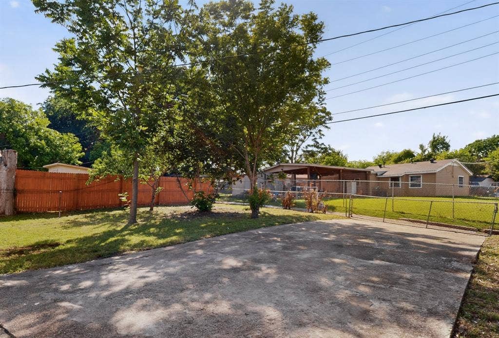 3853 Happy Canyon Drive, Dallas, TX 75241 Gated concrete driveway with a chain-link fence, mature trees, and a wooden privacy fence bordering a grass lawn