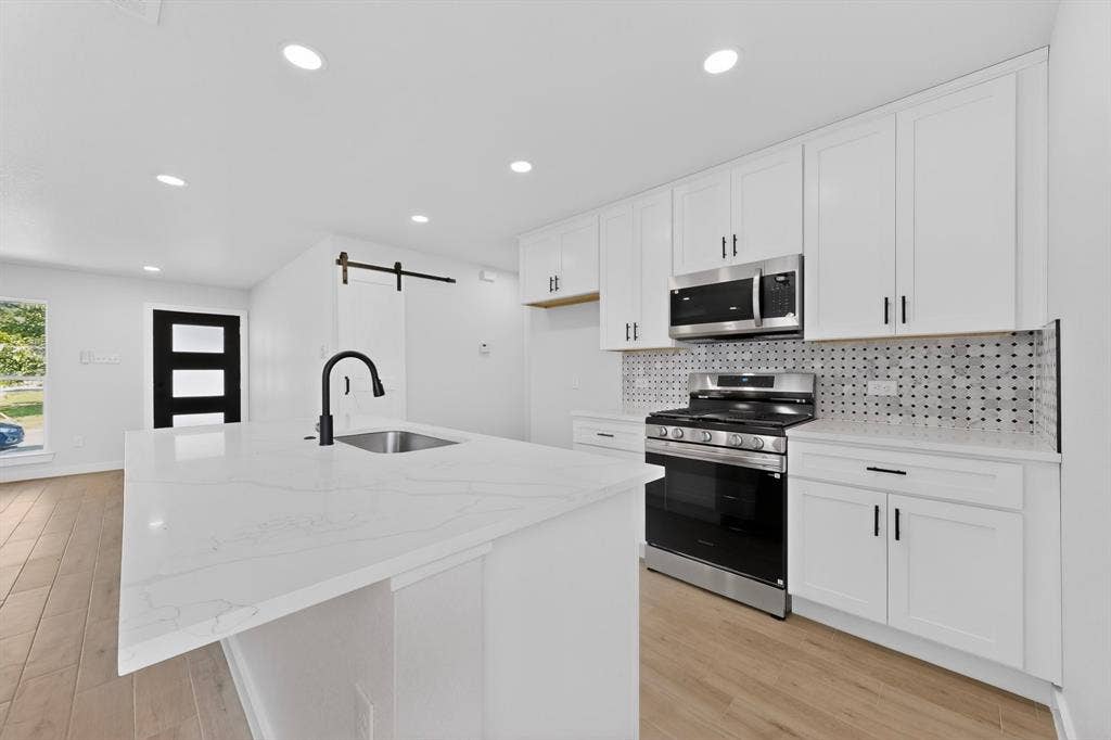 3853 Happy Canyon Drive, Dallas, TX 75241 Modern kitchen featuring an expansive island with an undermount sink and matte black faucet, stainless steel appliances, white cabinetry with contrasting hardware, and a graphic tile backsplash