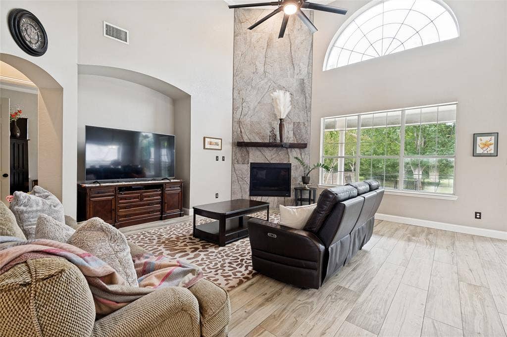 108 Appalachian Way, McKinney, TX 75071 Vaulted living room featuring a stone fireplace with a wood mantel, a ceiling fan, and large windows with an arched transom