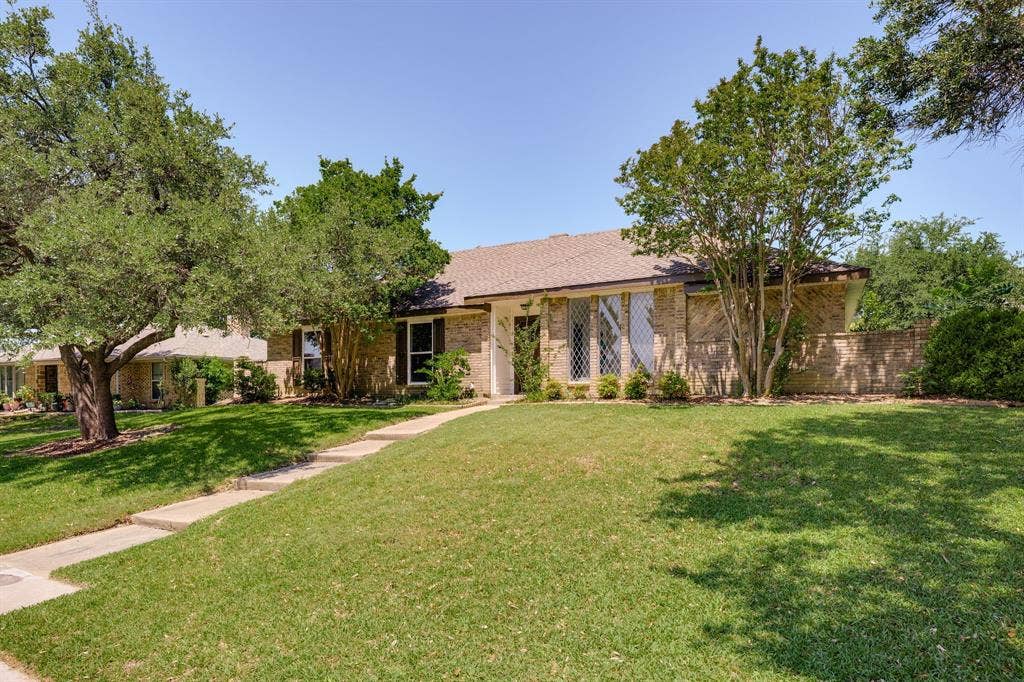 3406 Windridge Drive, Garland, TX 75043 Brick facade residence featuring a front lawn with mature trees and a concrete pathway