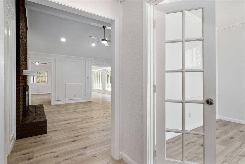 3406 Windridge Drive, Garland, TX 75043 French doors open to an expansive living area featuring wood-finish flooring, a brick fireplace, a vaulted ceiling, recessed lighting, and decorative wall paneling
