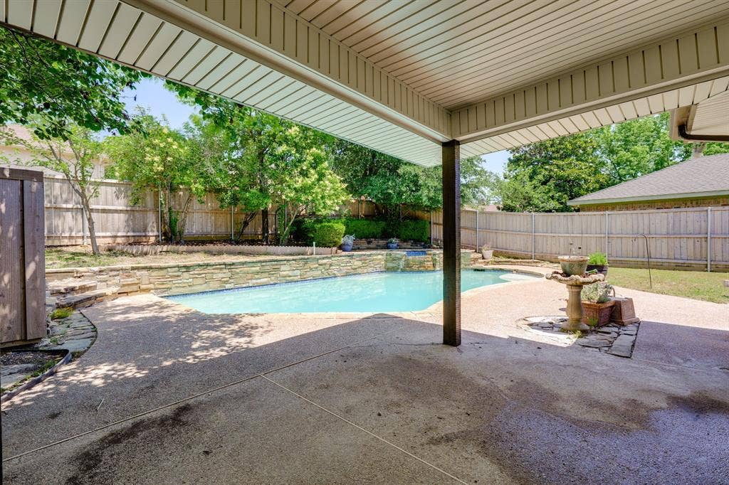 3406 Windridge Drive, Garland, TX 75043 Covered patio featuring an exposed aggregate concrete surface, leading to an inground pool with a stone coping and a waterfall feature