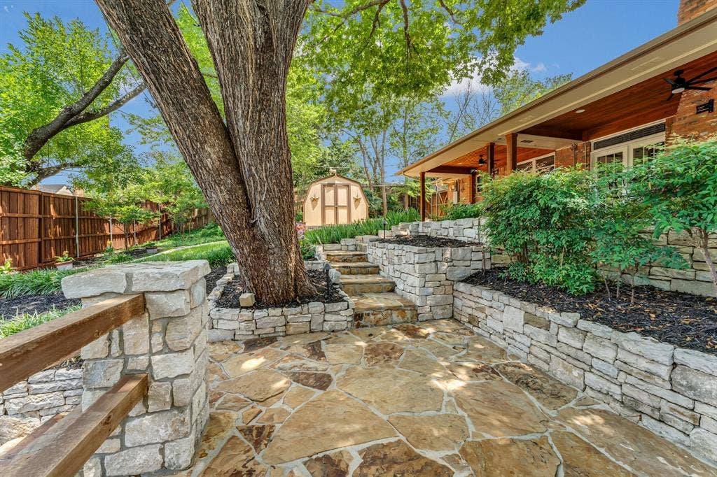 3908 Wildwood, Euless, TX 76040 Terraced backyard featuring flagstone patio, stone retaining walls, mature tree, wooden privacy fence, and covered patio with wood ceiling