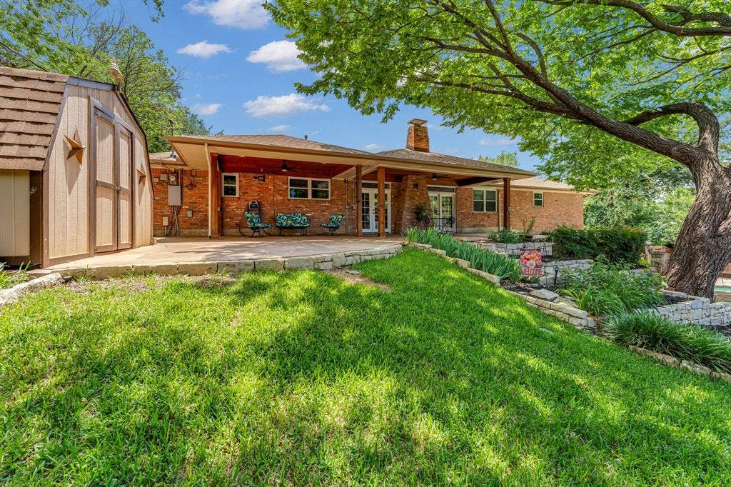 3908 Wildwood, Euless, TX 76040 Expansive backyard featuring a brick exterior home, covered patio, stone retaining walls, mature trees, and a separate storage shed with a shingled roof