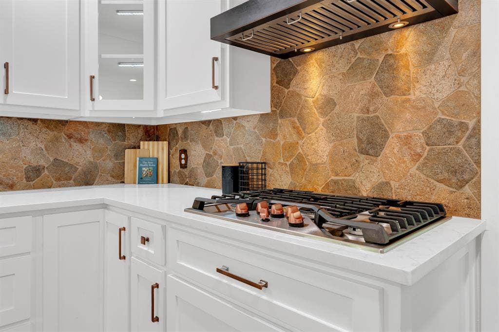3908 Wildwood, Euless, TX 76040 Built-in gas cooktop with coordinating range hood, stone slab backsplash, white cabinetry with bronze hardware, and light-colored countertops