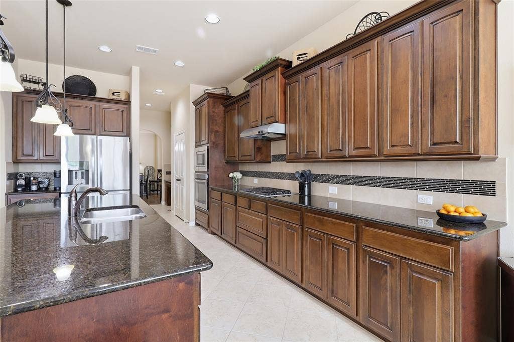 6905 Denali Drive, McKinney, TX 75070 Spacious kitchen featuring extensive dark wood cabinetry, granite countertops, stainless steel appliances, a tile backsplash with decorative accent, and recessed lighting