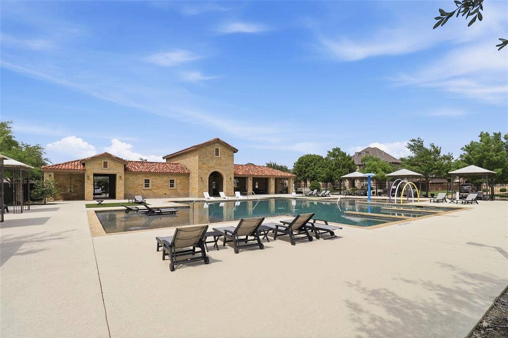6905 Denali Drive, McKinney, TX 75070 Resort-style pool area featuring a stone-clad building with a tile roof, multiple chaise lounges, a splash pad with water features, and shaded seating areas