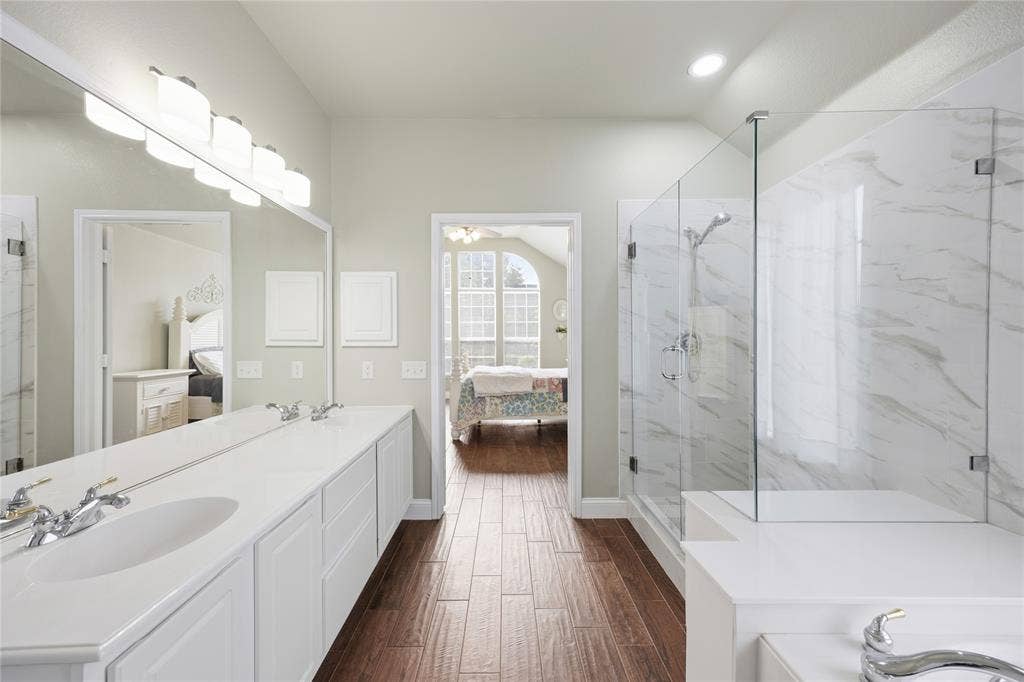 Wood-finish flooring throughout, featuring a dual-sink vanity with white cabinetry, a full-width mirror, and a glass-enclosed shower with white marble-patterned tile surround