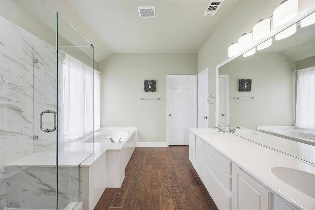 Spacious bathroom featuring a glass-enclosed shower with marble-look tile, a built-in soaking tub, a long double vanity with integrated sinks, and wood-finish flooring