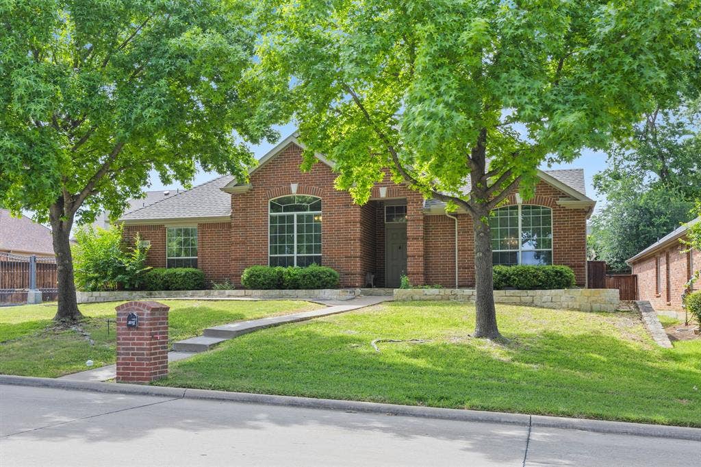 Traditional brick exterior featuring an arched window, a gabled roofline, mature trees, a manicured lawn, and a stone retaining wall