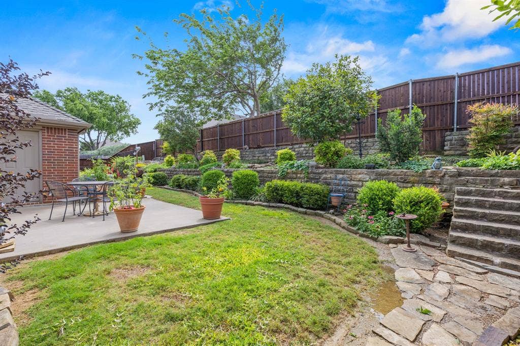 Expansive backyard featuring a stone patio, brick exterior, wood fence, tiered landscaping with stone retaining walls, and stone steps