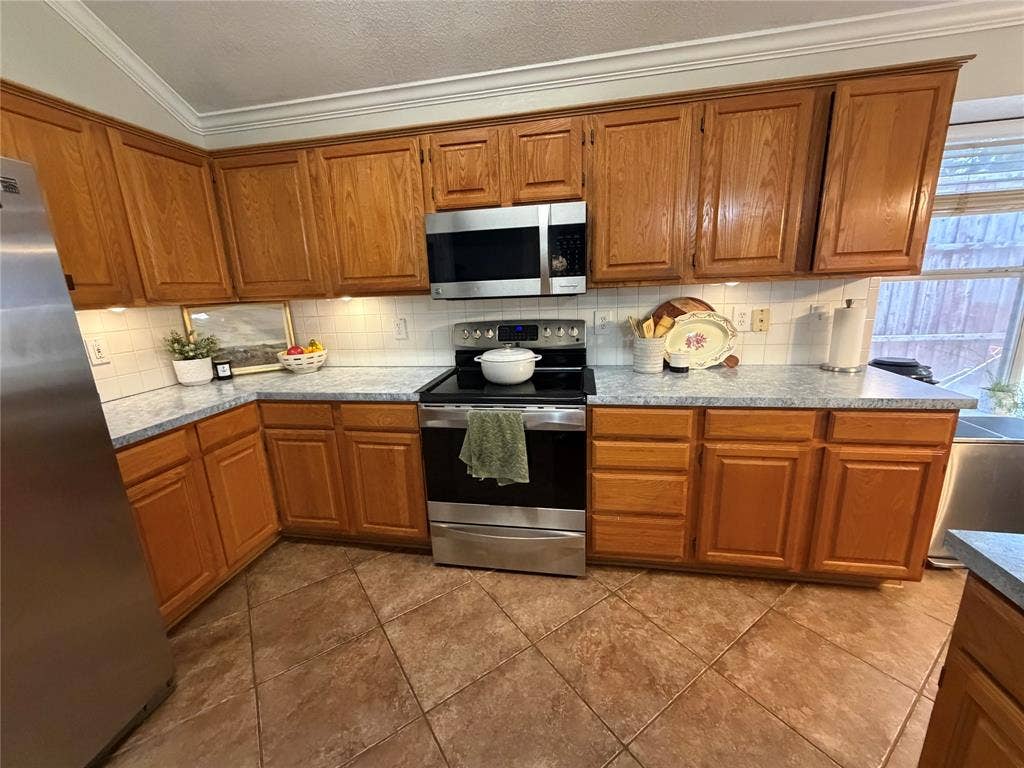 Wood-finish cabinetry, stainless steel appliances, tiled flooring, white subway tile backsplash, and crown molding