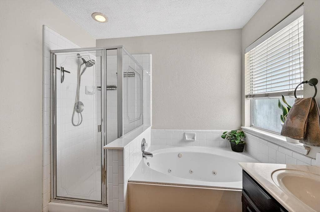Bathroom featuring a glass-enclosed shower, a white jetted soaking tub, a single window with blinds, a vanity with an undermount sink, and recessed lighting