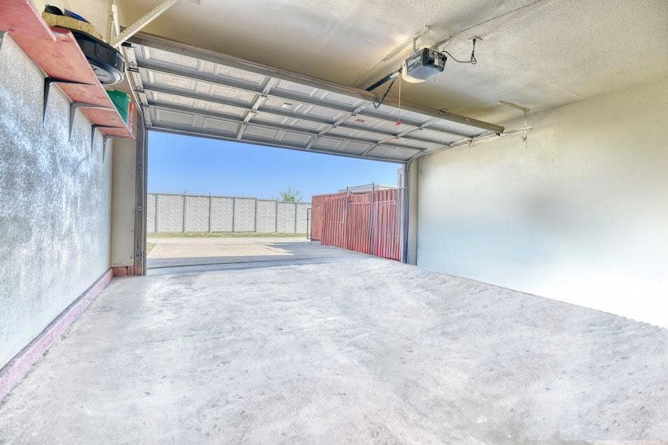 Concrete floor garage featuring an automatic garage door opener, overhead shelving, and a perimeter fence