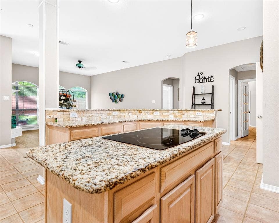 6005 Pin Oak Drive, McKinney, TX 75072 Kitchen island featuring a built-in electric cooktop, granite countertops, light wood-finish cabinetry, and a stone-tiled backsplash