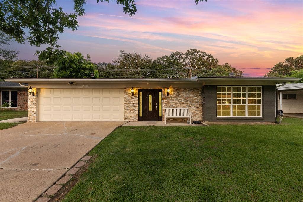 Mid-century ranch-style exterior featuring a two-car garage, stone veneer accents, a dark wood entry door, and a large multi-pane window