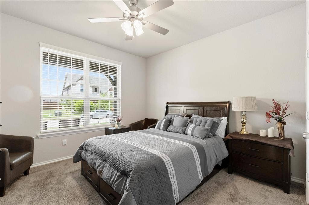 702 Matador Lane, Arlington, TX 76002 Bright bedroom featuring a large window with white blinds, neutral wall tones, textured carpeting, and a ceiling fan with integrated lighting