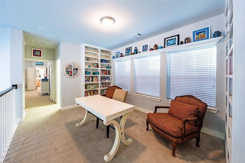 Carpeted upper-level library/den featuring extensive built-in shelving, three windows with blinds, and a flush-mount ceiling light fixture and custom made, art shelf about windows.