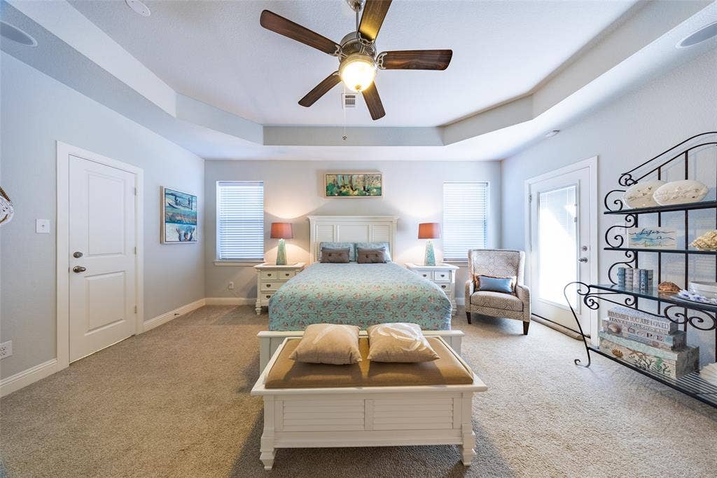 Carpeted Primary bedroom featuring a tray ceiling, ceiling fan with light fixture, and light gray wall paint and accessories - jewelry valet closet on left.  Door leading to private balcony to the right.